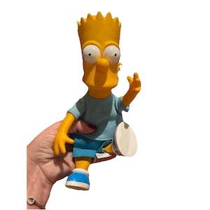 May include: A yellow Bart Simpson figurine with a spiky head, large eyes, and a mischievous grin. He wears a light blue t-shirt, blue shorts, and blue and white sneakers. The figurine is posed with one arm raised.
