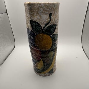 May include: A tall, textured ceramic vase with a beige speckled background. The vase is adorned with a colorful fruit design, including an orange, grapes, and pears. The fruit is painted in shades of yellow, green, purple, and red.