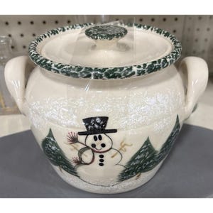 May include: Cream-colored ceramic pot with a lid, featuring a snowman and evergreen tree design. The pot has two handles and a green-trimmed rim. The snowman has a black top hat and a red scarf. The pot is likely for kitchen use.