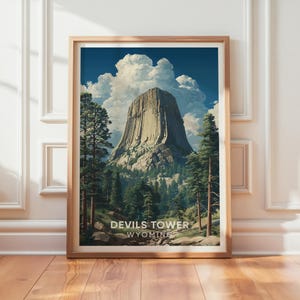 May include: A framed art print depicting Devils Tower in Wyoming. The image shows the distinctive rock formation set against a blue sky with white clouds. Green trees surround the base, with the text "Devils Tower Wyoming" at the bottom.