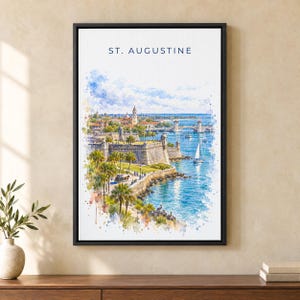 May include: A framed watercolor print titled ST. AUGUSTINE featuring a coastal cityscape with a stone fortress, palm trees, a bridge, and a sailboat on blue water. The artwork uses blue, green, and tan tones with a white border in a black frame.