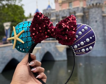 May include: A pair of black mouse ears with a red velvet bow, a blue and green mermaid scale ear with a yellow starfish, and a purple and gold seashell ear with white pearls.