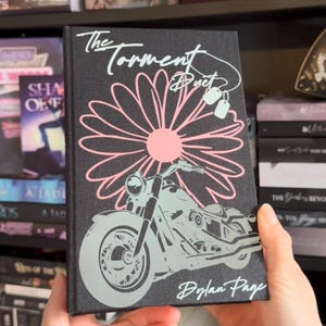 May include: A hardback book titled "The Torment Duet" by Dylan Page. The cover displays a light blue motorcycle graphic, a pink daisy, and the title in a stylized font. The book is held in front of a bookshelf.