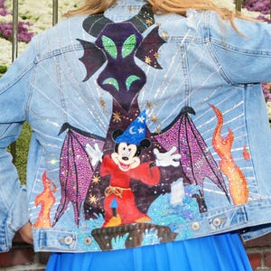 Puede incluir: Chaqueta vaquera azul claro con un gráfico colorido de un personaje de Disney y un dragón. La chaqueta se lleva delante de un letrero que dice "70 Celebrate Happy". El fondo incluye un edificio y una exhibición floral.