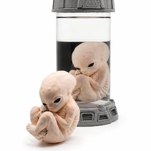 May include: A pale, fetal-like alien figure sits curled up next to a clear cylindrical container. Inside the container, a similar alien figure is suspended in a dark liquid. The container has a gray, industrial-style lid with a warning symbol.
