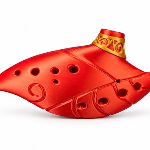 May include: A bright red ocarina shaped like a leaf with a gold-colored mouthpiece featuring embossed scrollwork. The instrument has multiple finger holes arranged across its surface and a subtle spiral design etched into the side of the body.