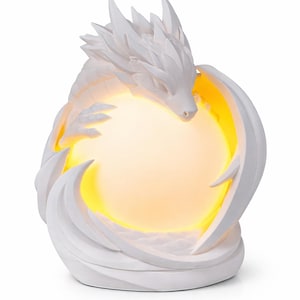 May include: A white dragon-shaped lamp with a glowing yellow orb at its center. The dragon is intricately detailed, with sharp features and wings that curve around the light source. The lamp is a decorative item.