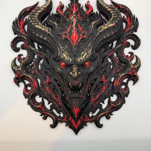 May include: A wall-mounted plaque featuring a sculpted demon head with large curved horns, glowing red eyes, and an open mouth showing fangs. The piece is primarily black with intricate red and gold painted highlights and ornate filigree detailing.