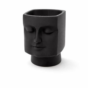 May include: A black, square-shaped vase with a sculpted face design. The face has closed eyes and a serene expression. The vase sits on a small, round base and is made of a matte material.