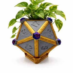 May include: A unique, geometric planter shaped like a twenty-sided die, with gold accents and purple gem-like details. The gray planter holds a vibrant green plant with yellow-green leaves. A decorative piece for home decor.
