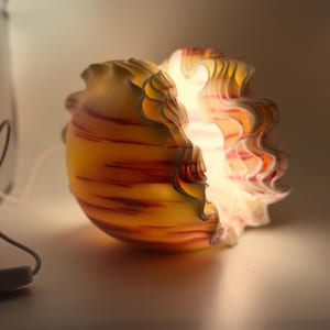 May include: A decorative glass lamp with a unique, layered design. The lamp features a yellow and red striped exterior with a ruffled edge. The interior glows with a warm light, creating a soft ambiance.