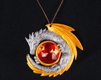 May include: A silver dragon pendant with gold wings and accents, encircling a large, faceted red gemstone. The pendant hangs from a brown cord necklace, set against a black background.