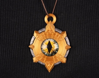 May include: A gold-colored pendant necklace with a detailed eye design. The pendant features a central eye with yellow and blue hues, surrounded by a gold and silver-colored frame. The necklace is suspended from a brown cord.