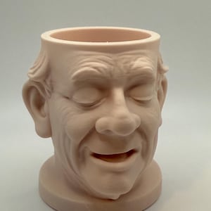 May include: A light pink, 3D-printed planter shaped like a smiling man's face. The planter has a round base and a wide opening at the top. The face has detailed wrinkles and closed eyes.