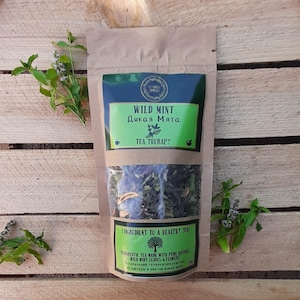 Pode incluir: Um saco de papel castanho de chá de hortelã selvagem com rótulos verdes e azuis. O rótulo superior diz "WILD MINT" em inglês e cirílico. Raminhos de hortelã fresca são dispostos ao redor do saco sobre uma superfície de madeira.