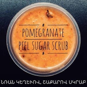 May include: A jar of pomegranate peel sugar scrub. The scrub has a grainy, orange appearance and is contained in a clear plastic jar. The text "POMEGRANATE PEEL SUGAR SCRUB" is printed on the jar. Below the text is text in a different language.