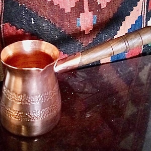 May include: A copper coffee pot with a wooden handle. The pot has a hammered texture and is filled with a dark liquid. The handle is attached to the side of the pot and is made of polished wood. The pot is sitting on a dark surface.