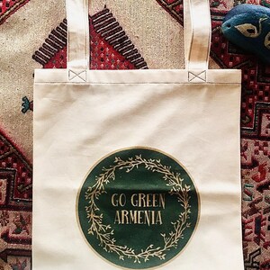 May include: Cream-colored tote bag with the text "GO GREEN ARMENIA" inside a green circle with a gold wreath design. The bag has cream-colored handles and is sitting on a patterned rug.