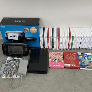 Puede incluir: Una consola Wii U negra con un mando, juegos y accesorios. La consola está en su caja original, con una pila de cartuchos de juego y varias carcasas de juegos. La caja es azul con el logotipo de Wii U.
