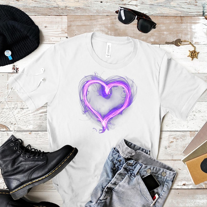 May include: White t-shirt with a purple heart design. The heart has a glowing, smoky effect. The image also shows blue jeans, black boots, and sunglasses. The shirt is made of a soft material.