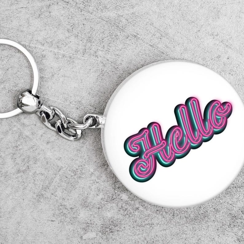 May include: A white circular keyring featuring the word "Hello" in a neon-style font. The word is pink with a teal outline. The keyring has a silver metal ring and chain.