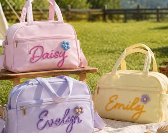 May include: Three rectangular duffel bags in pastel pink, light purple, and pale yellow. Each bag features a front zippered pocket, two top handles, and a detachable shoulder strap. The names Daisy, Evelyn, and Emily are embroidered in cursive.