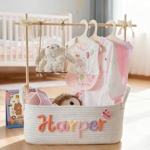 May include: A baby clothes rack with a white rope basket labeled "Harper" in colorful letters. The rack holds a plush bunny on a swing, baby clothes, and a bib with a bear design. A crib is in the background.