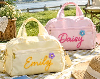 May include: A light yellow duffel bag with the name Emily embroidered in yellow and a purple flower patch, and a light pink duffel bag with the name Daisy embroidered in pink and a blue flower patch, both featuring top handles and zippers.