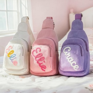 May include: Three corduroy sling bags in white, pink, and purple. Each bag has a front pocket and a name embroidered on it: "Ellie" in colorful thread, "Ellie" in pink, and "Grace" in white. The bags are on a bed.