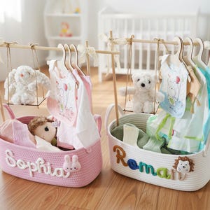 May include: Two decorative baby clothes organizers. One pink basket with the name "Sophia" and a bunny, and one white basket with the name "Roman" and a lion. Each has a wooden swing with a stuffed animal and baby clothes.
