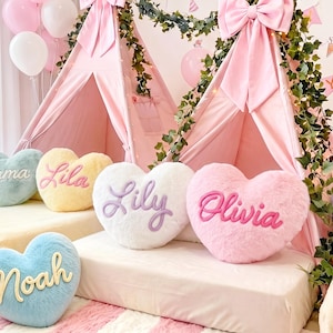 May include: A collection of plush heart-shaped cushions in various pastel colours, including mint green, yellow, white, pink, blue, and purple. Each cushion features a name embroidered in cursive script: Emma, Lila, Lily, Olivia, Noah, and Sophia.