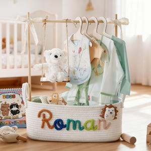 May include: A white rope basket with the name "Roman" in colorful letters, holding baby clothes and toys. A teddy bear swings from a wooden rack above, with a calendar and wooden blocks nearby. The clothes are light green and white.