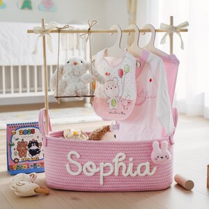 May include: A pink rope basket with the name "Sophia" in white script, holding baby clothes on hangers. A plush bunny swings from a wooden frame above. A 2023 calendar and wooden toys are nearby.