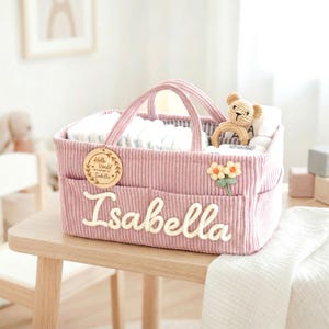 May include: Pink corduroy baby basket with handles, pockets, and the name "Isabella" in white script. The basket contains diapers, a teddy bear, and a flower applique. A wooden tag reads "Hello World, My Name is Isabella."