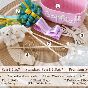 May include: A pink woven basket with the name "Sophia" in white script, surrounded by various craft supplies. Includes wooden dowel rods, hangers, plush dolls, a mini swing, plastic bags, and blue and pink pull-bow ribbon.