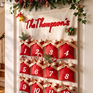 Può includere: Un calendario dell'avvento in tessuto color crema appeso a una porta, con tasche rosse numerate da uno a dodici. Ogni tasca è decorata con un fiocco di nastro dorato e il testo The Thompson's è ricamato in filo rosso nella parte superiore.