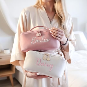 May include: Two cosmetic bags are held in front of a person. The top bag is pink with the name "Emilia" embroidered in white. The bottom bag is off-white with the name "Daisy" embroidered in pink. Both bags have gold zippers.