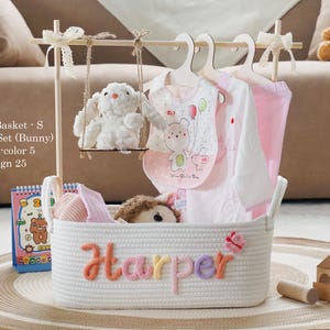 Op de afbeelding: Een witte touwmand met de naam "Harper" in kleurrijke letters. De mand bevat babykleding, een pluchen konijn op een schommel en een slab. De tekst "White Basket - S Premium Set (Bunny) Muliti-color 5 Design 25" is zichtbaar.
