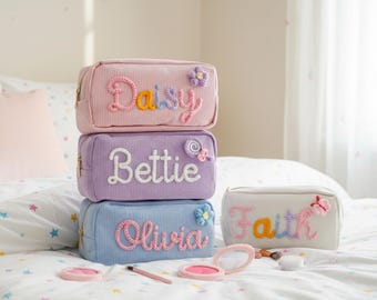 May include: A stack of four corduroy cosmetic bags in pastel colors: pink, lavender, and blue. Each bag has a name embroidered in a contrasting color: Daisy, Bettie, Olivia, and Faith. Decorative flowers and butterflies adorn the bags.