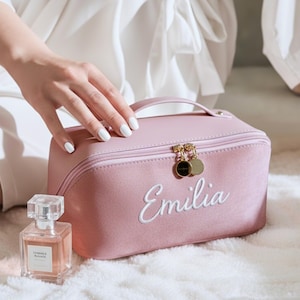 Może przedstawiać: Różowa kosmetyczka z białym haftem z imieniem "Emilia". Torba ma uchwyt i złote suwaki. Obok torby stoi butelka perfum.
