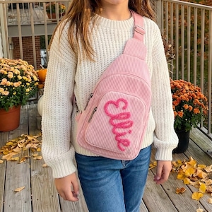 Peut inclure: Un sac bandoulière en velours côtelé rose avec le nom "Ellie" brodé sur le devant. Le sac a une seule sangle et une poche zippée. La personne porte un pull blanc et un jean bleu.