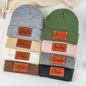 May include: A selection of personalised beanies in shades like grey, green, beige, pink, white, and brown. Each beanie has a brown leather patch with a name engraved. Names include Paula, Rachel, Justin, Emily, Sandra, Sophia, Jackson, and Olivia. A cosy winter accessory.
