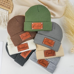 Puede incluir: Una colección de gorros personalizados en varios colores, incluyendo verde, marrón, gris y crema. Cada gorro presenta un parche rectangular de cuero con un nombre grabado. Los nombres incluyen Rachel, Justin, Sandra, Paula, Emily, Olivia y Jack.