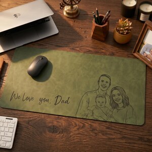 Könnte beinhalten: Eine rechteckige olivgrüne Schreibtischunterlage mit einer schwarzen Strichzeichnung einer Familie, bestehend aus zwei Erwachsenen und einem Baby. Der Text "We love you, Dad" ist in Schreibschrift auf der linken Seite der Oberfläche gedruckt.
