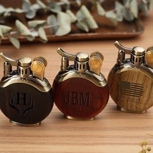 May include: Three personalized lighters with a round design. The lighters have a brass finish with wooden and dark brown accents. One lighter has the letter "H" engraved, another has the initials "JBM", and the third has an American flag.