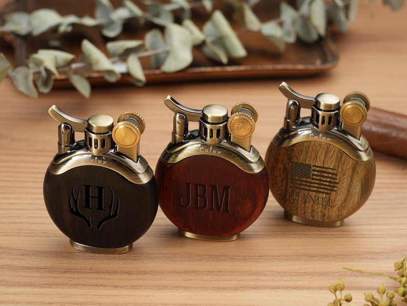 May include: Three personalized lighters with a round design. The lighters have a brass finish with wooden and dark brown accents. One lighter has the letter "H" engraved, another has the initials "JBM", and the third has an American flag.