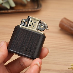 May include: A dark wood-grain lighter with a brass-colored top. The lighter has the initials "MC" engraved on the front. The lighter is held in a person's hand.