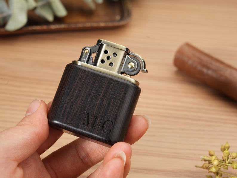May include: A dark wood-grain lighter with a brass-colored top. The lighter has the initials "MC" engraved on the front. The lighter is held in a person's hand.