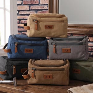 Può includere: Una collezione di sei beauty case rettangolari in tela color beige, blu, grigio, grigio scuro e verde oliva. Ogni borsa presenta una tasca frontale con cerniera, una maniglia laterale e una toppa in pelle marrone con iniziali o nomi impressi.