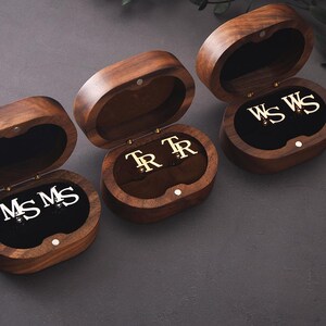 May include: Three open wooden jewellery boxes, each displaying a pair of initialled cufflinks. The boxes are oval, with a dark brown finish and a black velvet lining. The cufflinks feature silver or gold initials. The boxes are open.
